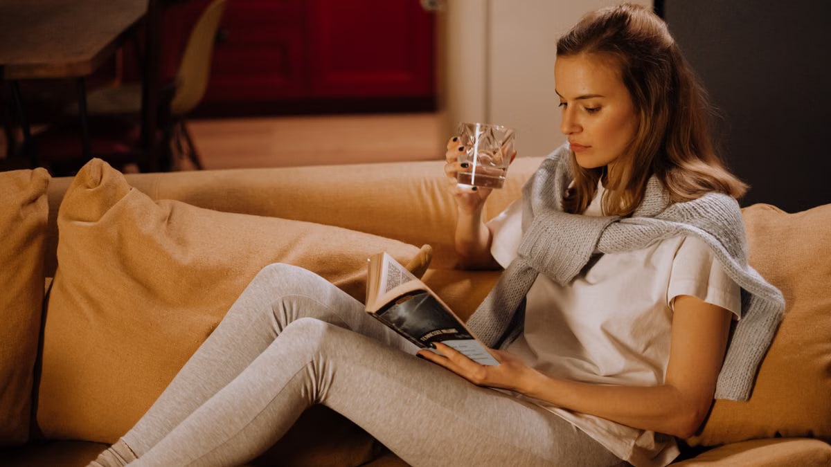 Woman relaxing on sofa, reading a book and drinking water in a warm, inviting living room setting.